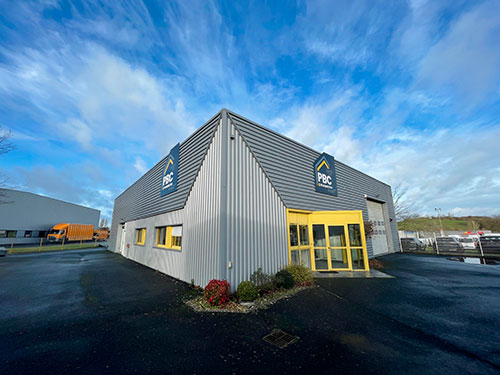 Bâtiment actuel de PBC Plaquistes à Cholet, avec bardage métallique gris, entrée vitrée jaune et enseignes PBC, sous un ciel bleu nuageux.