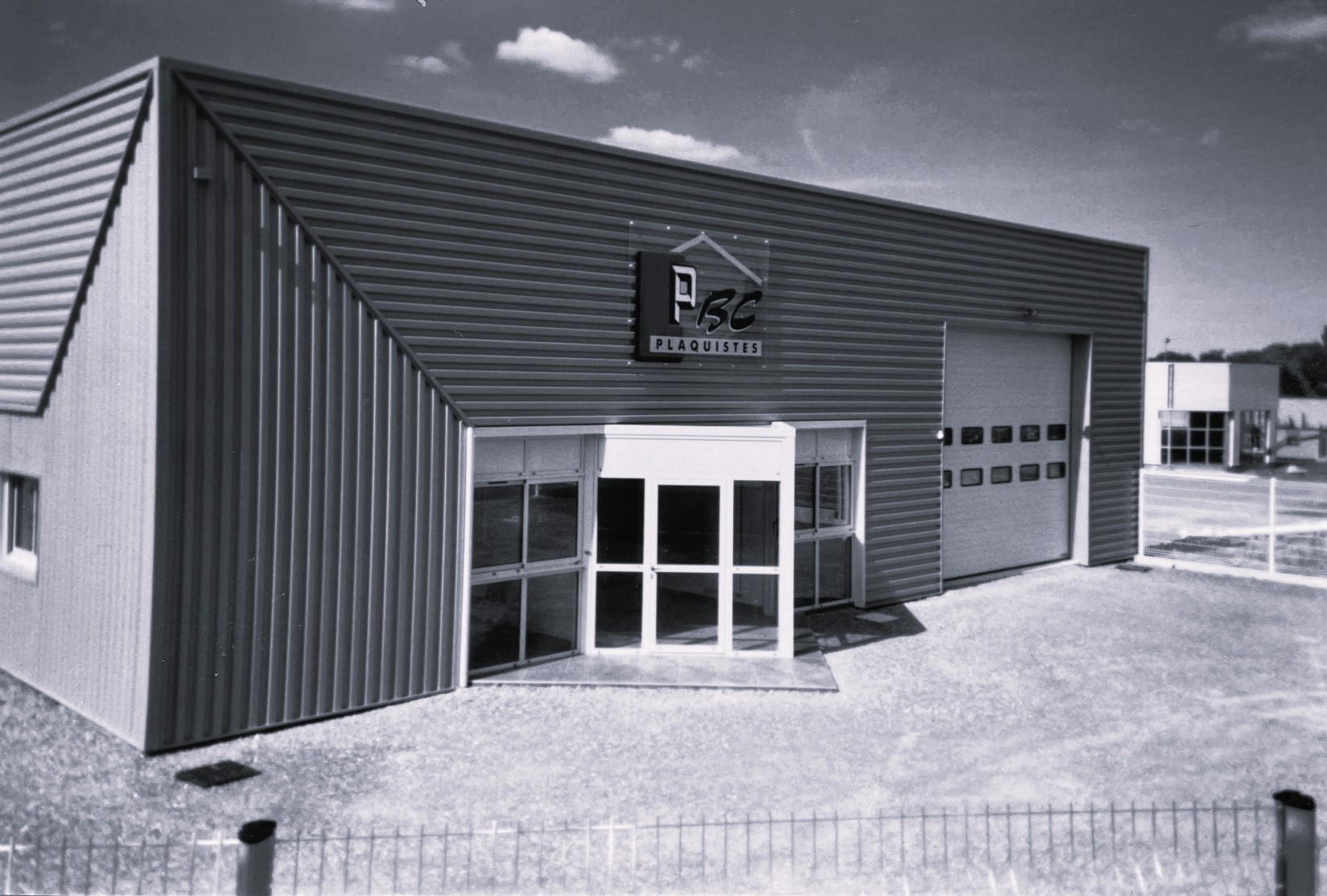 Ancien bâtiment PBC Plaquistes en noir et blanc, avec bardage métallique, entrée vitrée centrale et porte industrielle, illustrant l’histoire et les débuts de l’entreprise.