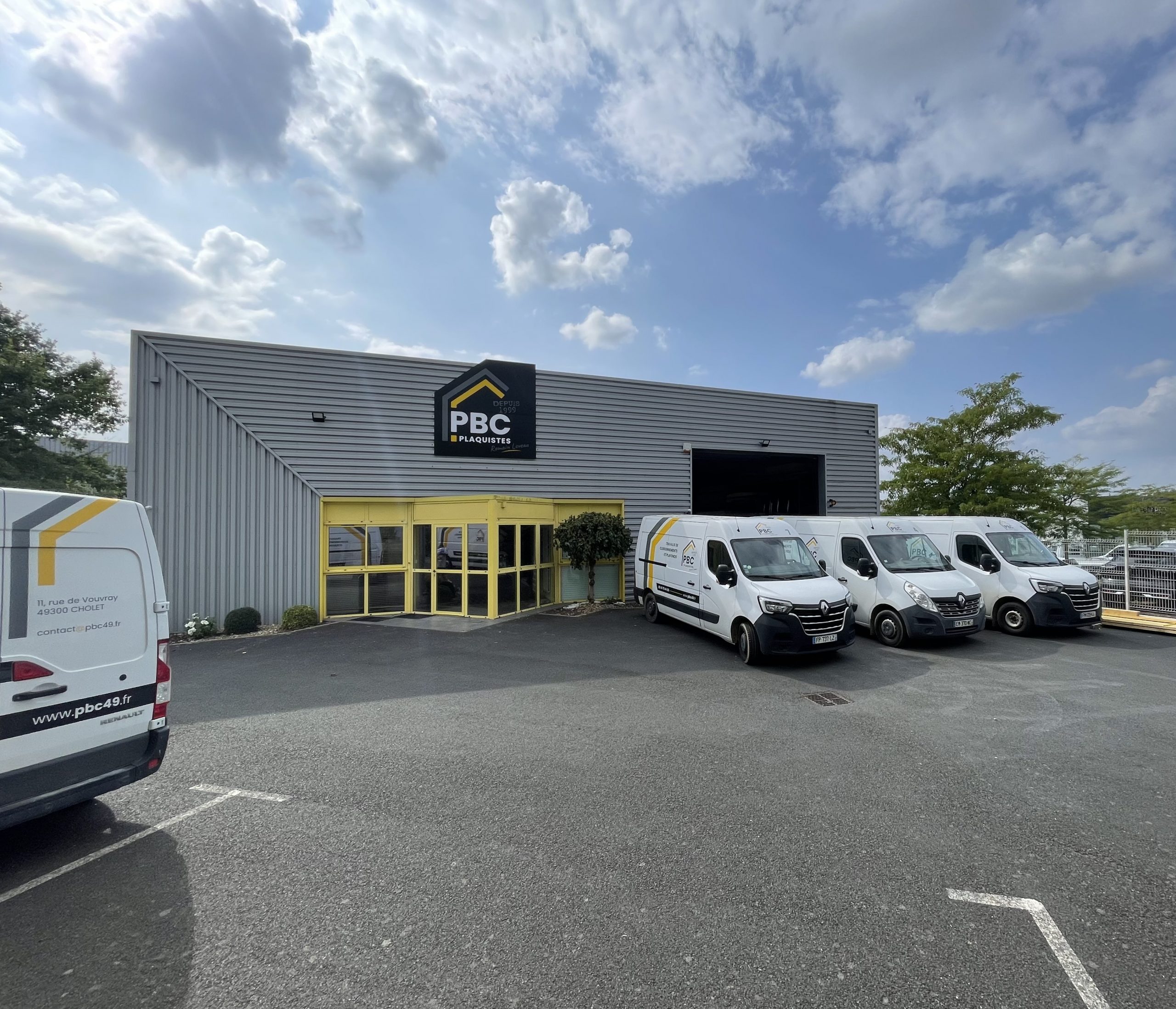 Façade du bâtiment PBC Plaquistes à Cholet, avec bardage métallique gris, entrée vitrée jaune et véhicules utilitaires de l’entreprise stationnés devant.